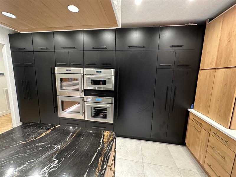 Matte black floor-to-ceiling cabinet wall with built-in ovens beside natural wood cabinets